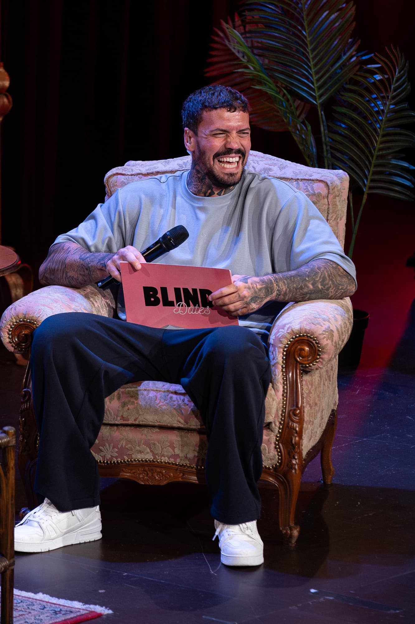 EMMVEE lacht herzhaft im Sessel auf der Blind-Dates-Bühne, die Moderationskarte in der Hand