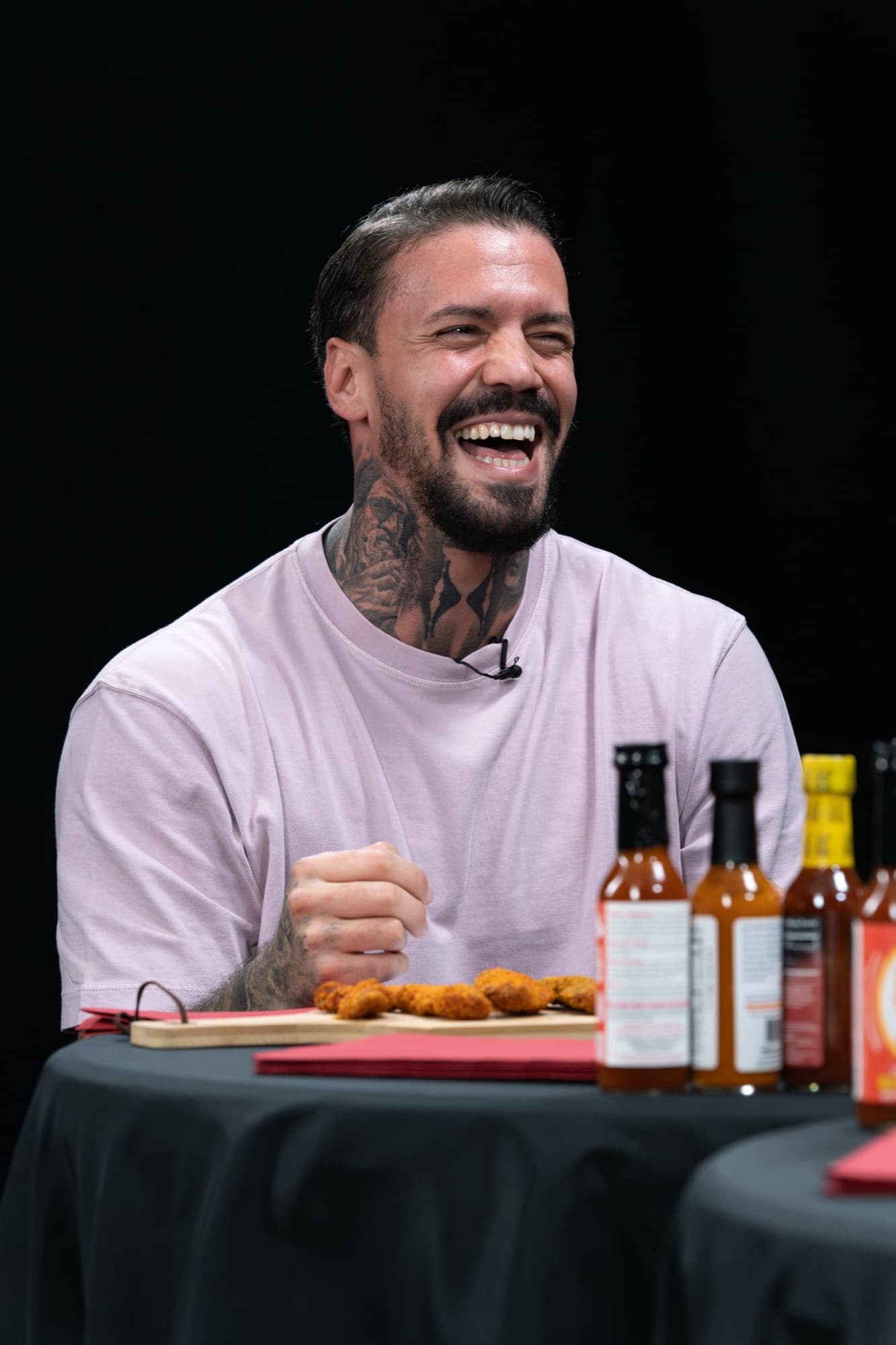 EMMVEE lacht bei Hot Ones Germany, vor ihm die Reihe Hot-Sauce-Flaschen